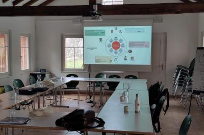 A meeting room with tables arranged in a U-shape and a projector screen showing a slide titled "Swiss DBGI-KM team" with team members and work packages.